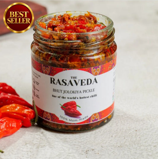 A glass jar of Rasaveda Bhut Jolokiya Pickle (one of the world's hottest chili pickles) on a light textured surface with fresh red chili peppers and a gold 'Best Seller' badge overlay.