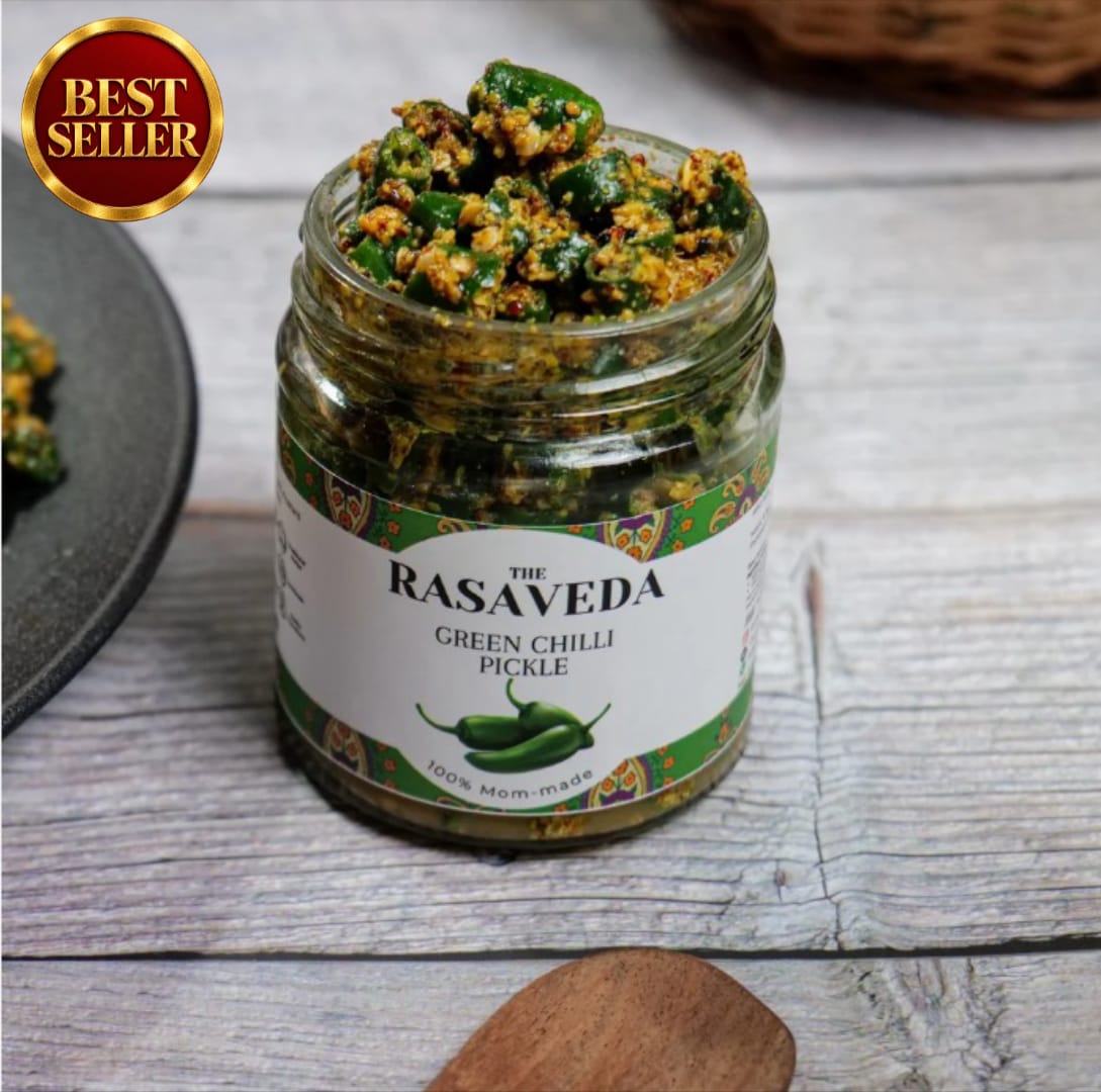 An open glass jar of The Rasaveda Green Chilli Pickle filled with chopped green chilies and spices, set on a rustic gray wooden surface with a wooden spoon and a dark plate partially visible.
