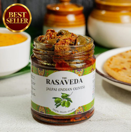 A jar of Rasaveda Jalpai (Indian Olives) on a light surface with a 'BEST SELLER' badge in the top left. The jar is filled with green olives in a reddish-brown sauce, with blurred bowls of food in the background.