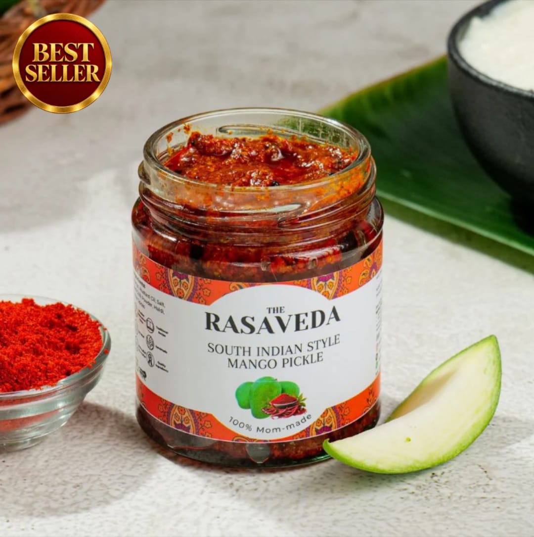 A jar of Rasaveda South Indian Style Mango Pickle on a light textured surface, styled with a slice of mango, a small bowl of red mango powder, and a green leaf in the background. A gold 'Best Seller' badge is in the top-left corner.